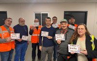Eine Gruppe von neun Personen steht in einem Innenraum und hält jeweils ein kleines, bedrucktes Blatt Papier hoch. Einige Personen tragen orange Warnwesten oder Arbeitskleidung. Im Hintergrund sind helle Wände, Türen und ein Fenster zu sehen.