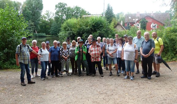 Gruppe von etwa 25 Personen auf einem breiten Weg am Ufer eines kleinen Gewässers. Einige tragen Rucksäcke, Hüte oder Wanderstöcke. Im Hintergrund stehen dichte Bäume und Sträucher sowie ein rotes Gebäude mit der Aufschrift „Restaurant“. Die Szene befindet sich im Freien bei Tageslicht.