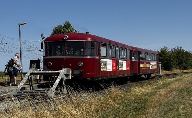 Roter zweiteiliger Schienenbus steht auf einem Gleis an einem Prellbock. Links stehen mehrere Personen auf einem Bahnsteig. Im Vordergrund befinden sich trockenes Gras und Schotter, im Hintergrund Oberleitungen, Bäume und blauer Himmel. Auf der Fahrzeugseite sind mehrere Plakate und Schilder angebracht.