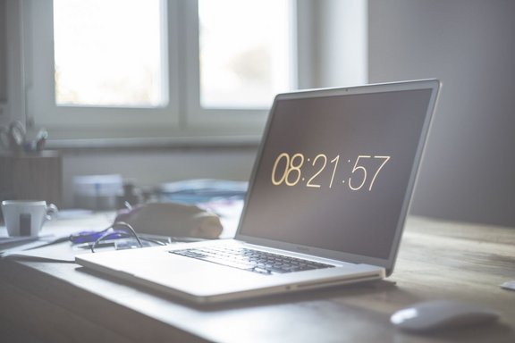 Laptop auf einem Holztisch in einem hellen Raum. Auf dem Bildschirm ist die Uhrzeit „08:21:57“ zu sehen. Neben dem Laptop liegen eine Brille, ein Etui oder eine Tasche und weitere unscharfe Gegenstände. Links steht eine weiße Tasse. Im Hintergrund befinden sich große Fenster, die viel Tageslicht hereinlassen. Die Aufnahme ist insgesamt leicht unscharf mit hellem, weichem Licht.