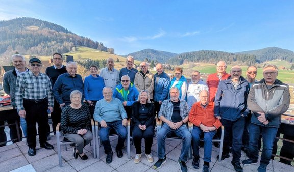 Gruppenfoto von 22 Personen, die auf einer Terrasse im Freien posieren. Vorne sitzen fünf Personen auf Stühlen, dahinter stehen die übrigen in zwei Reihen. Im Hintergrund sind grüne Wiesen, einzelne Gebäude und bewaldete Hügel unter blauem Himmel zu sehen.