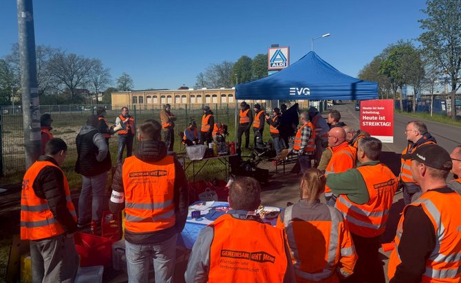 Gruppe von Personen im Freien auf einem befestigten Platz, viele tragen orange Warnwesten. In der Mitte stehen Tische mit Getränken und Gegenständen. Dahinter ein blaues Faltzelt mit der Aufschrift „EVG“. Rechts neben dem Zelt ein rotes Schild mit weißer Schrift: „Heute ist STREIKTAG!“. Im Hintergrund ein Zaun, Bäume, ein Gebäude. Himmel klar und blau.