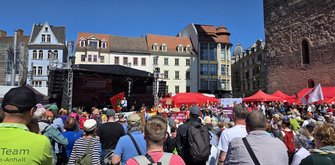 Große Menschenmenge auf einem sonnigen Stadtplatz vor einer Bühne mit schwarzem Dach und Lautsprecheranlagen. Im Vordergrund stehen dicht gedrängt viele Personen, einige mit Rucksäcken und Kopfbedeckungen. Auf der Bühne spricht eine Person ins Mikrofon; davor sind Fahnen und Banner in Rot und Gelb zu sehen. Rund um den Platz stehen mehrstöckige historische Gebäude mit unterschiedlichen Fassaden und Dächern. Der Himmel ist klar und blau.