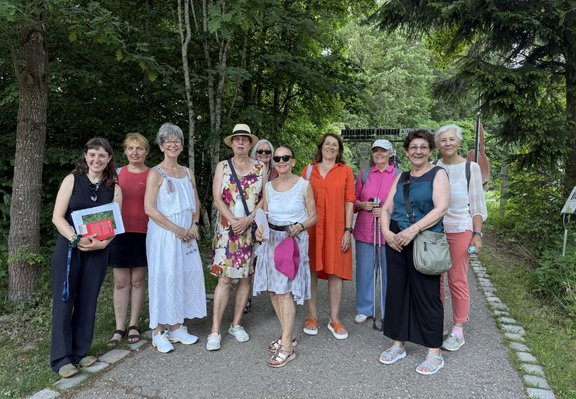 Neun Personen stehen nebeneinander auf einem asphaltierten Weg in einer grünen Park- oder Gartenanlage. Im Hintergrund sind dichtes Laub, Bäume und ein Tor oder Eingang zu sehen. Die Personen tragen sommerliche Kleidung in verschiedenen Farben, darunter Weiß, Rot, Orange, Pink, Blau und Schwarz. Eine Person hält ein Buch oder eine Mappe, eine andere zwei Gehstöcke. Mehrere Personen tragen Taschen oder Rucksäcke. Der Weg ist von Gras und Pflanzen gesäumt, das Licht wirkt natürlich und gleichmäßig. Geben Sie Ihr Feedback zu BizChat ab.