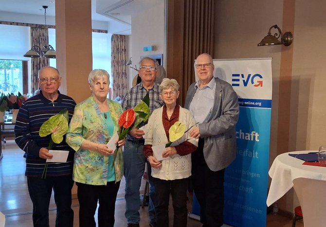 Fünf Personen stehen in einem Innenraum vor einem Roll-up mit dem EVG-Logo. Vier Personen halten jeweils eine Blüte und eine weiße Karte in den Händen. Im Hintergrund sind Vorhänge, Wandlampen und Fenster sichtbar.