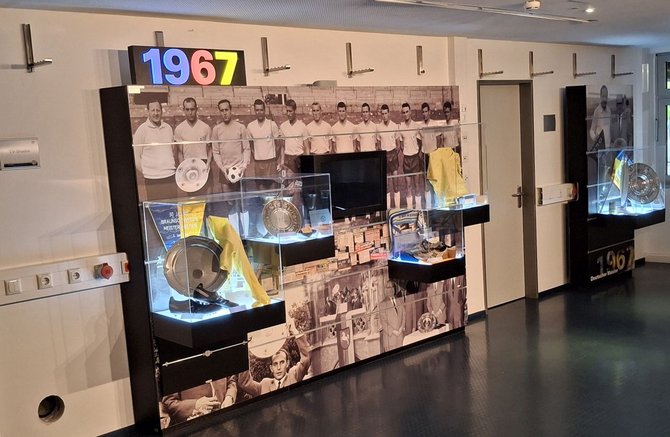 Ein Ausstellungsbereich mit der großen Zahl „1967“ zeigt eine historische schwarz-weiße Mannschaftsfotografie und mehrere beleuchtete Glasvitrinen mit Pokalen, Tellerplatten, Dokumenten und einem gelben Trikot. Die Exponate sind entlang einer Wand angeordnet, der Boden ist dunkel.