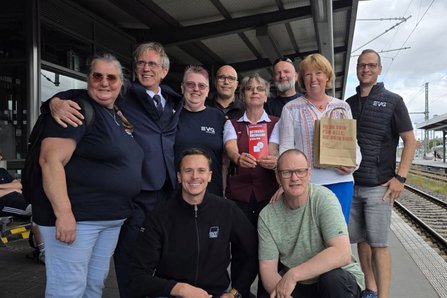 Eine Gruppe von Menschen steht auf einem Bahnsteig neben den Gleisen und posiert dicht zusammen für ein Foto. Einige tragen dunkle Kleidung mit Aufdruck; eine Person hält ein rotes Heft, eine andere eine Papiertüte. Zwei Personen sitzen vorne, die übrigen stehen dahinter.