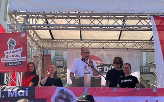 Bühne im Freien mit Metallträger-Dach. In der Mitte steht ein Rednerpult mit Mikrofon, dahinter ein Banner. Auf einem Schild am Pult steht: „Erst unsere Jobs, dann eure Profite – 1. Mai 2026“. Links hängt ein rotes Plakat mit erhobener Faust und demselben Text sowie dem Kürzel „DGB“. Mehrere Personen stehen auf der Bühne neben und hinter dem Rednerpult. An der Bühnenfront hängt ein rotes Banner mit der Aufschrift „1. Mai“.