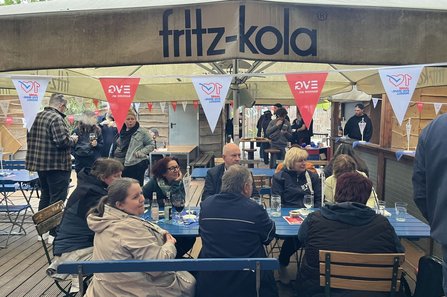 Außenbereich mit Holzboden und mehreren blauen Biertischen und -bänken, an denen zahlreiche Personen sitzen und stehen. Über den Tischen sind große beige Sonnenschirme mit der Aufschrift „fritz‑kola“ gespannt. Daran hängen rot‑weiße Wimpel mit Texten wie „EVG kommt an.“ und „10 Jahre EVG‑Wahlmodell“ sowie einem Herzsymbol. Auf den Tischen stehen Gläser, Flaschen und kleine Fähnchen. Im Hintergrund sind weitere Stehtische, Personen und Holzwände zu sehen.