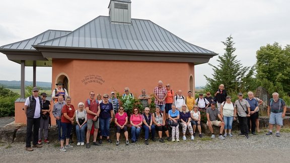 Eine große Gruppe von etwa 25–30 Personen macht ein Gruppenfoto vor einer kleinen orangefarbenen Kapelle in einer ländlichen Umgebung. Die Personen stehen und sitzen in mehreren Reihen vor der Kapelle. Einige sitzen auf einer niedrigen Mauer, andere stehen dahinter oder daneben. Viele tragen Freizeitkleidung und Wanderschuhe, einige haben Rucksäcke und Hüte. Die Kapelle hat ein graues Metalldach mit einem kleinen Turmaufsatz. Links befindet sich ein überdachter Anbau. Im Hintergrund sind Felder, Bäume und eine leicht hügelige Landschaft zu sehen.