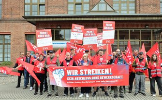Gruppe von Demonstrierenden vor einem Backsteingebäude mit zahlreichen roten EVG-Fahnen. Mehrere Personen halten Schilder mit der Überschrift „MERKT EUCH DAS:“. Vor der Gruppe wird ein großes Banner gehalten mit der Aufschrift: „WIR STREIKEN! KEIN ABSCHLUSS OHNE GEWERKSCHAFT @GBM: Unsere Forderungen sind kein Hexenwerk!“.