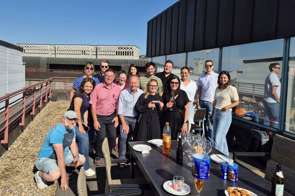 Gruppenfoto auf einer Dachterrasse bei sonnigem Wetter: Etwa 14 Personen stehen und sitzen um einen dunklen Tisch mit Gläsern, Flaschen, Schalen und Tellern. Im Vordergrund kniet eine Person neben dem Tisch. Rechts befindet sich eine Glasfassade, die Teile der Gruppe spiegelt. Im Hintergrund sind weitere Gebäudeteile, Geländer und ein klarer blauer Himmel zu sehen.