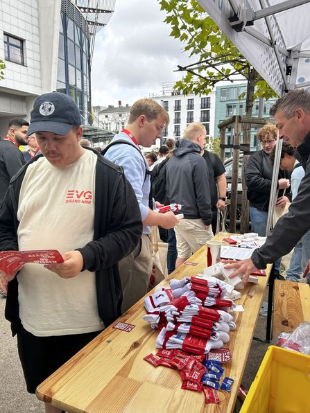 Außenbereich unter einem Pavillon. Auf einem langen Holztisch liegen gefaltete weiße Textilien mit roten Etiketten, rote Flyer, kleine verpackte Gegenstände, Kartenstapel und Becher. Mehrere Personen stehen am Tisch und halten Informationsmaterial oder Stofftaschen. Im Hintergrund sind weitere Personen, Gebäude, Bäume und ein bewölkter Himmel sichtbar.
