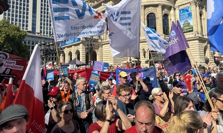 Dicht gedrängte Menschenmenge auf einem Platz vor einem historischen Gebäude mit Säulen und Fahnenmasten. Mehrere weiße EVG-Fahnen und weitere Gewerkschaftsfahnen ragen über die Menge. Sichtbare Schilder tragen unter anderem die Aufschriften „Hart verdient!“, „Ja zum Sozialstaat“ und „Teilhabe ohne Barrieren“. Im Hintergrund sind moderne Hochhäuser und Banner am Gebäude zu sehen.