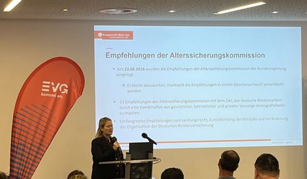 Ein Vortragsraum mit Sitzpublikum. Links steht ein rotes EVG-Rollbanner mit der Aufschrift „EVG kommt an.“. Vor einer Projektionsfläche spricht eine Person an einem Rednerpult mit Mikrofon. Auf der Leinwand ist eine Präsentationsfolie mit dem Titel „Empfehlungen der Alterssicherungskommission“ zu sehen. Sichtbarer Text umfasst unter anderem „Knappschaft Bahn See“ sowie mehrere Stichpunkte zur Alterssicherungskommission.
