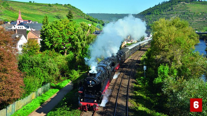Schwarze Dampflokomotive mit mehreren Personenwagen auf einer zweigleisigen Bahnstrecke. Weiße Dampfwolken steigen über den Zug auf. Die Strecke verläuft durch eine grüne Tallandschaft mit Bäumen, Häusern und Weinbergen an den Hängen. Rechts ist ein Fluss zu sehen. Unten rechts befindet sich eine rote Markierung mit der Zahl „6“.