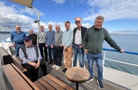 Gruppe mehrerer Personen auf dem offenen Deck eines Boots. Eine Person sitzt links im Vordergrund auf einer Holzbank, trägt eine Kappe, ein helles Hemd und eine dunkle Weste. Mehrere weitere Personen stehen nebeneinander auf dem Holzdeck, überwiegend in langen Hosen, Pullovern oder Jacken in Blau‑, Grau‑, Grün‑ und Beigetönen; eine Person lehnt rechts an einem blauen Geländer. In der Mitte des Decks steht ein kleiner runder Holztisch. Links sind weitere Holzbänke und ein senkrechter Metallmast mit Sonnensegel zu sehen. Im Hintergrund erstreckt sich eine Wasserfläche mit leichtem Wellengang, dahinter eine entfernte Uferlinie. Darüber ein heller Himmel mit weißen Wolken und blauem Himmel.