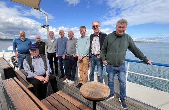 Gruppe mehrerer Personen auf dem offenen Deck eines Boots. Eine Person sitzt links im Vordergrund auf einer Holzbank, trägt eine Kappe, ein helles Hemd und eine dunkle Weste. Mehrere weitere Personen stehen nebeneinander auf dem Holzdeck, überwiegend in langen Hosen, Pullovern oder Jacken in Blau‑, Grau‑, Grün‑ und Beigetönen; eine Person lehnt rechts an einem blauen Geländer. In der Mitte des Decks steht ein kleiner runder Holztisch. Links sind weitere Holzbänke und ein senkrechter Metallmast mit Sonnensegel zu sehen. Im Hintergrund erstreckt sich eine Wasserfläche mit leichtem Wellengang, dahinter eine entfernte Uferlinie. Darüber ein heller Himmel mit weißen Wolken und blauem Himmel.