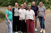 Sieben Personen stehen auf einem gepflasterten Weg im Freien vor dichtem grünen Baumbestand. Vier Personen stehen in der vorderen Reihe und drei Personen dahinter. Die Personen tragen überwiegend Freizeitkleidung in verschiedenen Farben, darunter gestreifte Oberteile, ein türkisfarbenes T‑Shirt, eine weiße Bluse, eine dunkle kurze Hose und helle Schuhe. Im Hintergrund sind Bäume, Sträucher und eine niedrige Betonbegrenzung zu sehen. Auf einem dunklen Oberteil ist der Schriftzug „GANT“ erkennbar.