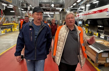 Mehrere Personen gehen durch eine Werkhalle neben einem weißen Hochgeschwindigkeitszug mit rotem Streifen. Eine Person trägt eine orangefarbene Warnweste, eine andere eine dunkelblaue Arbeitsjacke mit DB-Logo. In der Halle sind Arbeitsplätze, Regale, technische Anlagen und Holzpaletten sichtbar. Der Boden ist teilweise rot markiert und die Halle ist hell beleuchtet.