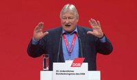 Eine Person steht hinter einem Rednerpult vor einem vollständig roten Hintergrund. Auf dem Pult sind zwei Mikrofone befestigt, links daneben steht ein Glas mit Wasser. Die Person trägt ein dunkles Jackett über einem hellen Hemd und ein rotes Schlüsselband mit Ausweis um den Hals. Beide Hände sind vor dem Oberkörper erhoben, die Handflächen zeigen nach außen. Auf der Vorderseite des Rednerpults ist ein rotes Logo mit der Aufschrift „DGB“ sowie darunter der Text „23. Ordentlicher Bundeskongress des DGB“ zu sehen.
