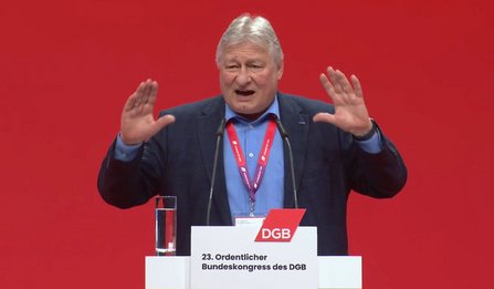 Eine Person steht hinter einem Rednerpult vor einem vollständig roten Hintergrund. Auf dem Pult sind zwei Mikrofone befestigt, links daneben steht ein Glas mit Wasser. Die Person trägt ein dunkles Jackett über einem hellen Hemd und ein rotes Schlüsselband mit Ausweis um den Hals. Beide Hände sind vor dem Oberkörper erhoben, die Handflächen zeigen nach außen. Auf der Vorderseite des Rednerpults ist ein rotes Logo mit der Aufschrift „DGB“ sowie darunter der Text „23. Ordentlicher Bundeskongress des DGB“ zu sehen.