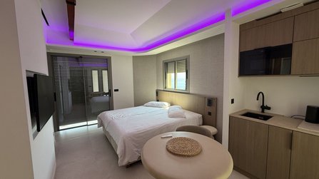 Wohnraum mit Doppelbett, rundem Tisch, Küchenzeile und Wandfernseher. Rechts befindet sich eine Küchenzeile mit Spüle und Oberschränken. Eine große Glasfront mit teilweisem Rollladen trennt den Raum von einem Balkonbereich. Violette indirekte Beleuchtung verläuft entlang der Decke.