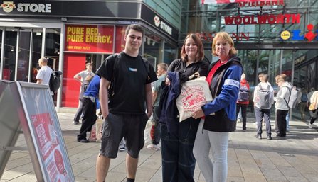 Vor einem Geschäft mit der Aufschrift „STORE“ stehen drei Personen auf einer Fußgängerfläche. Im Hintergrund sind weitere Personen sowie die Schriftzüge „PURE RED. PURE ENERGY.“, „WOOLWORTH“ und Logos von Geschäften sichtbar. Eine Person hält eine helle Stofftasche mit rotem Aufdruck. Links im Vordergrund steht ein Werbeaufsteller.