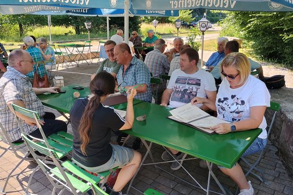Mehrere Personen sitzen an grünen Tischen in einem Biergarten unter blauen Sonnenschirmen und betrachten Speisekarten oder unterhalten sich. Im Vordergrund sitzen mehrere Personen an zwei zusammengestellten grünen Tischen. Eine Person hält eine Brille in der Hand, eine andere blättert in einer Speisekarte. Auf den Tischen liegen Speisekarten, Mobiltelefone und kleine Gegenstände. Die Sitzgelegenheiten bestehen aus zusammenklappbaren grünen Metallstühlen.