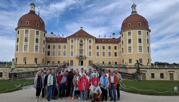 Etwa 35 Personen stehen und sitzen vor einem großen barocken Schloss mit zwei runden Ecktürmen und roten Kuppeldächern. Vor dem Gebäude führt eine breite Treppe zu einer Terrasse mit Balustraden und mehreren Statuen. Im Vordergrund befinden sich Kieswege und Rasenflächen mit Absperrketten. Der Himmel ist überwiegend blau mit verstreuten Wolken.