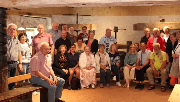Eine größere Gruppe von Personen sitzt und steht gemeinsam in einem rustikalen Raum und posiert für ein Gruppenfoto. Die Personen sind in zwei Reihen angeordnet: Einige sitzen vorne auf Holzbänken, andere stehen dahinter. Der Raum hat grobe Wände, Holzbalken und alte Geräte oder Pressen aus Holz und Metall, die an einer Wand befestigt sind. Der Boden besteht aus rötlichen Fliesen. Die Personen tragen unterschiedliche Alltagskleidung und schauen in Richtung Kamera. Einzelne Taschen und persönliche Gegenstände liegen am Boden oder auf den Bänken.
