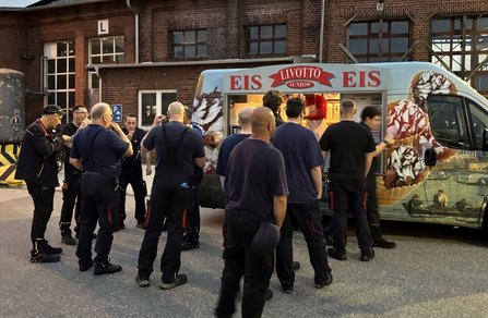 Mehrere Personen in dunkler Arbeitskleidung stehen vor einem Eisverkaufswagen. Auf dem Fahrzeug ist der Schriftzug „LIVOTTO JUNIOR EIS“ zu sehen. Im Hintergrund befindet sich ein Backsteingebäude mit großen Fenstern und einem Tor mit dem Buchstaben „L“. Einige Personen halten Eis in der Hand oder stehen in einer Warteschlange vor dem Verkaufsfenster.