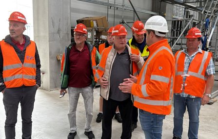 Mehrere Personen stehen in einem Rohbau- oder Technikbereich mit Betonwänden und Gerüstteilen. Alle tragen Schutzhelme, die meisten zusätzlich orangefarbene Warnwesten. Eine Person mit weißem Helm spricht mit der Gruppe. Im Hintergrund stehen Materialbehälter und Bauelemente.