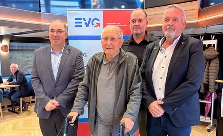 Vier Personen stehen in einem Innenraum vor einem Roll-up-Banner mit der Aufschrift „EVG Eisenbahn- und Verkehrsgewerkschaft“. Eine Person in der Mitte hält einen Rollator. Im Hintergrund sind Tische, Sitzgelegenheiten und eine Garderobe mit aufgehängten Jacken zu sehen. Der Raum hat einen hellen Holzfußboden und große Fensterflächen.