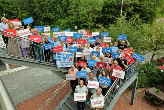Große Gruppe von Menschen steht im Freien und hält vor sich oder über den Kopf zahlreiche rechteckige Schilder in den Farben Blau, Rot und Weiß. Auf den Schildern sind kurze Slogans in deutscher Sprache zu lesen, jeweils mit „EVG“ als Absender. Einige Personen halten zusätzlich eine blaue Fahne mit weißer Beschriftung. Im Hintergrund sind Pflanzen, ein Gebäude mit heller Fassade und gestapelte Holzscheite zu sehen. Die Personen sind unterschiedlich gekleidet, stehen dicht beieinander und blicken überwiegend zur Kamera.