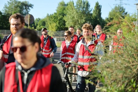 Mehrere Personen in roten Warnwesten bewegen sich auf einem Weg im Freien. Im Vordergrund sind unscharfe Personen zu sehen, in der Mitte schiebt eine Person ein Fahrrad. Im Hintergrund stehen Bäume, Sträucher und Verkehrsschilder.