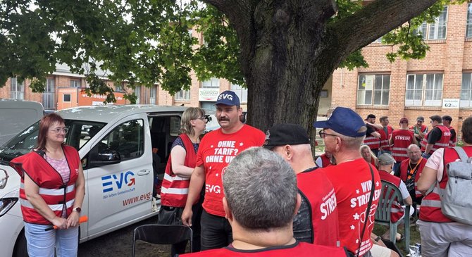 Mehrere Personen in roten EVG-Westen und roten T-Shirts stehen und sitzen unter einem großen Baum vor einem Backsteingebäude. Links steht ein weißes Fahrzeug mit EVG-Logo und der Aufschrift „Wir leben Gemeinschaft“ sowie der Internetadresse „www.evg-online.org“. Im Vordergrund stehen Stühle, im Hintergrund weitere Personen in roten Westen.