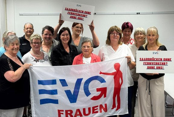 Elf Personen stehen in einem Innenraum vor einer weißen Wand und halten gemeinsam ein großes Banner mit dem EVG-Logo und dem Schriftzug „EVG FRAUEN“. Zwei Personen halten Plakate mit dem Text „SAARBRÜCKEN OHNE FERNVERKEHR? OHNE UNS!“. Die Gruppe ist in zwei Reihen angeordnet; das Banner befindet sich im Vordergrund. Hinter der Gruppe sind eine Wand mit Schienenleisten sowie mehrere Stühle am rechten Bildrand zu sehen.
