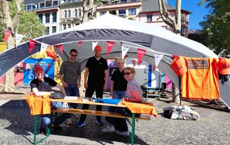 Infostand im Freien auf einem gepflasterten Platz. Ein großes graues Zelt mit Tischen und Bänken darunter. Mehrere Personen stehen oder sitzen am Tisch. Orangefarbene Westen, T-Shirts und Kappen mit „EVG“-Logo liegen auf dem Tisch oder hängen am Zelt. Im Hintergrund sind weitere Stände, Plakate und Gebäude zu sehen.