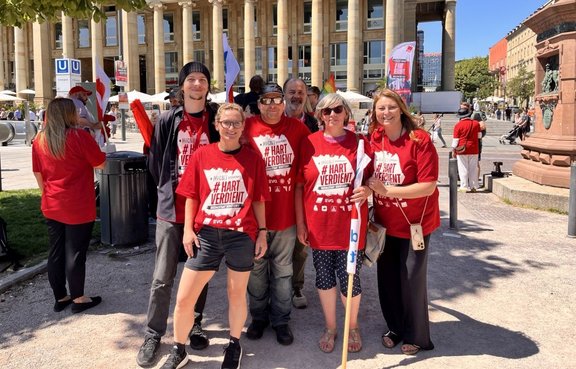 Gruppenfoto von sechs Personen im Vordergrund auf einem öffentlichen Platz vor einem Gebäude mit hoher Säulenfassade. Die Personen tragen überwiegend rote T‑Shirts mit dem gut lesbaren Aufdruck „# HART VERDIENT“. Einige halten Fahnen oder Fahnenstangen. Im Hintergrund sind weitere Personen, Fahnen, ein U‑Bahneingang mit „U“-Schildern, Sitzbereiche sowie ein Denkmal aus rotem Stein zu sehen. Sonniges Wetter mit starken Schatten auf dem Boden.