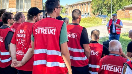 Mehrere Personen in roten Warnwesten mit der Aufschrift „STREIK!“ stehen im Freien und blicken zu einer sprechenden Person mit Mikrofon. Im Hintergrund sind Backsteingebäude, Grünflächen und geparkte Fahrzeuge zu sehen. Die Aufnahme entstand bei hellem Tageslicht.
