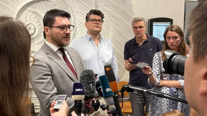 Eine Gruppe von Personen steht in einem Innenraum vor einer dekorativen Wand mit reliefartigem Muster. Eine Person in einem hellen Sakko spricht in mehrere Mikrofone, die von verschiedenen Aufnahmegeräten gehalten werden; auf einigen Mikrofonen sind Senderlogos wie „ZDF“ und „SWR“ sichtbar. Mehrere andere Personen halten Smartphones oder Aufnahmegeräte in Richtung der sprechenden Person. Im Hintergrund ist ein Monitor oder Display zu sehen.