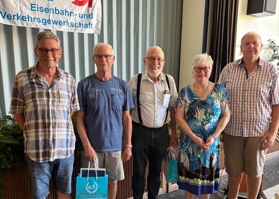 Fünf Personen stehen nebeneinander in einem Innenraum vor einem aufgehängten Banner mit der Aufschrift „EVG Eisenbahn- und Verkehrsgewerkschaft“. Vier Männer und eine Frau sind zu sehen, alle schauen in die Kamera. Zwei Personen halten Taschen, eine davon mit einer sichtbaren „65“-Aufschrift. Hinter ihnen sind Vorhänge und eine Pflanze erkennbar.
