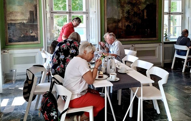 Mehrere Personen sitzen an weißen Tischen und Stühlen in einem hellen Raum mit großen Fenstern und großformatigen Wandgemälden. Auf den Tischen stehen Tassen, Teller, Gläser und eine Wasserkaraffe; einige Personen essen oder trinken. Im Vordergrund sitzt eine Person in roter Hose an einem gedeckten Tisch, daneben stehen Rucksäcke an den Stühlen.