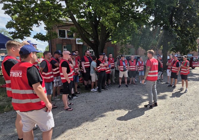 Eine Gruppe von Personen in roten EVG-Westen steht im Halbkreis auf einem Schotterplatz unter Bäumen. Im Zentrum spricht eine Person mit dem Rücken zur Kamera zu der Gruppe. Im Hintergrund sind Backsteingebäude, Fahrzeuge und weitere Personen zu sehen.