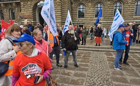 Mehrere Personen stehen auf einem Platz vor einem historischen Gebäude und halten weiße Fahnen mit dem Schriftzug „Eisenbahn- und Verkehrsgewerkschaft“. Im Vordergrund trägt eine Person eine blaue Kappe und ein rotes T-Shirt mit der Aufschrift „Hart verdient!“. Im Hintergrund sind weitere Teilnehmende, Fahnen und Schilder sichtbar.