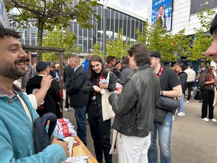 Außenbereich auf einem Platz mit Bäumen vor einer großen Glasfassade. Im Vordergrund steht ein Holztisch mit ausgelegten roten Flyern, gefalteten weißen Textilien und weiteren Werbematerialien. Mehrere Personen stehen um den Tisch herum, einige halten Stofftaschen mit rotem Aufdruck. Im Hintergrund sind weitere Besucherinnen und Besucher auf dem Platz verteilt.