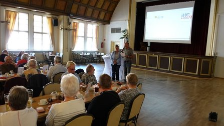 Großer Veranstaltungsraum mit mehreren langen Tischen und vielen sitzenden Personen. Eine Person steht mit Mikrofon vor einer Bühne und spricht in Richtung des Publikums. Auf der Bühne befindet sich eine große Projektionsleinwand. Sonnenlicht fällt durch die hohen Fenster auf der linken Seite in den Raum. Auf den Tischen stehen Getränke, Gläser und Tassen.