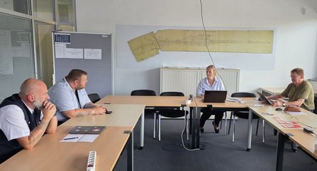 Vier Personen sitzen an einem rechteckig angeordneten Tisch in einem Besprechungsraum; auf dem Tisch stehen Laptops und Unterlagen, an der Wand hängt eine große Karte oder Zeichnung.