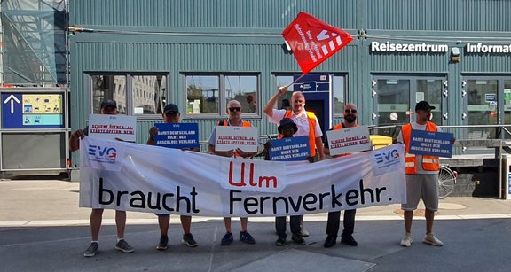 Sieben Personen stehen vor einem Gebäude mit der Beschriftung „Reisezentrum Information“. Die Personen tragen teilweise orange Warnwesten und halten ein großes weißes Banner mit der Aufschrift „Ulm braucht Fernverkehr.“ Mehrere Personen halten Schilder mit den Texten „Schiene öffnen! Ja. Städte opfern. Nein!“ und „Damit Deutschland nicht den Anschluss verliert.“ Eine Person hält eine rote Fahne mit dem ver.di-Logo. Im Hintergrund sind Fenster, Türen, Hinweisschilder und ein DB-Automat zu sehen.